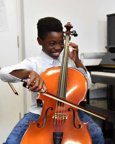 Two young students playing the cello together, with a piano behind them.